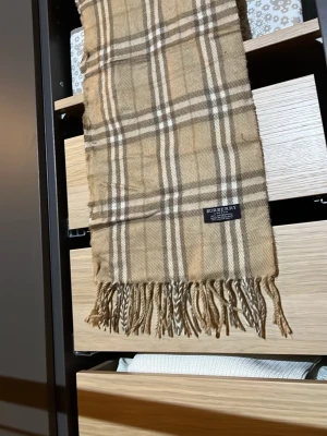 Beige rutig halsduk från Burberry - Klassisk beige halsduk från Burberry i 100% lammull med det ikoniska rutmönstret i brunt, vitt och svart. Halsduken har fransar i ändarna och är tillverkad i England. Perfekt accessoar för att lyfta din outfit.