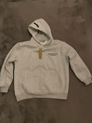 Grå Essentials Fear of God hoodie  - Snygg grå hoodie från Essentials Fear of God med svart logga på bröstet och ryggen. Klassisk magficka framtill, ribbade muddar och en stor huva. Perfekt för en avslappnad och trendig streetwear-look. Tveka inte vid frågor