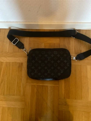 Louis Vuitton svart axelväska monogram - Snygg svart axelväska från Louis Vuitton med klassiskt monogrammönster i grått. Väskan har justerbar bred axelrem och silverfärgade metalldetaljer. Perfekt för dig som vill ha en stilren och lyxig accessoar till din outfit.
