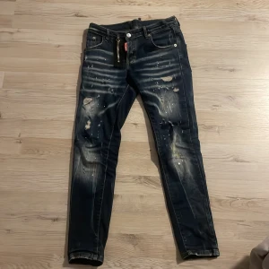 Dsquared2 jeans - Snygga mörkblå jeans med coola slitningar och färgstänk. De har en klassisk femficksdesign och knappgylf. Perfekta för en avslappnad stil med en edgy touch.  Storlek 152-164 