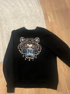 Svart Kenzo sweatshirt med tigertryck - Cool svart sweatshirt från Kenzo med det ikoniska tigerhuvudet broderat framtill och texten 'Kenzo Paris' under. Tröjan har rund halsringning och ribbade muddar vid ärmslut och nederkant. Perfekt för dig som gillar streetwear och statement-plagg.