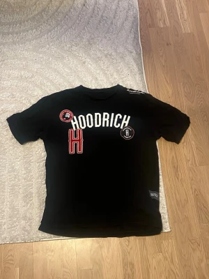 Svart Hoodrich t-shirt med tryck - Svart t-shirt från Hoodrich med flera tryck och patchar i vitt och rött framtill. Klassisk rund halsringning och korta ärmar. Perfekt för dig som gillar streetwear och vill sticka ut med en snygg logga.