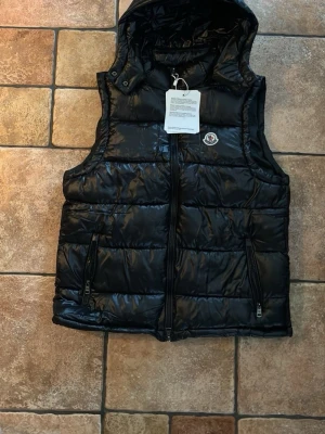 Svart dunväst från Moncler med huva - Snygg svart dunväst från Moncler med glansig finish, avtagbar huva och två dragkedjeförsedda fickor. Västen har Moncler-logga på bröstet och stängs med dragkedja framtill. Perfekt för lager på lager och streetwear-vibe.