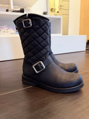 Svarta quiltade bikerboots med spännen - Snygga svarta bikerboots med quiltat skaft och två silvriga spännen. Bootsens nedre del är i slätt läder medan skaftet har ett vadderat rutmönster. Rund tå och låg klack ger en cool och robust look. Perfekta för dig som vill ha en edgy stil.
