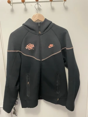 Svart Nike hoodie med dragkedja - Svart hoodie från Nike med dragkedja framtill och två fickor med dragkedja. Orange Nike-logga och grafiskt tryck på bröstet. Kontrastfärgade sömmar i beige. Tillverkad i mjukt material som känns skönt mot huden. Perfekt för en sportig och avslappnad stil.