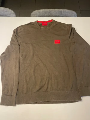 Mörk grön tröja från HUGO med röd logga - Snygg lite mörkare grön tröja från HUGO med rund halsringning och en tydlig röd logga på bröstet. Tröjan har ribbade muddar vid ärmslut och nederkant. Perfekt för dig som gillar stilrena plagg med en twist.