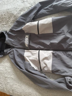 Hoodrich vindjacka - Snygg vindjacka från Hoodrich i grått med vita paneler och svarta dragkedjor. Jackan har huva, två stora fickor framtill och Hoodrich-logga på bröstet. Tillverkad i lätt och vindtätt material, perfekt för streetwear och chill dagar.