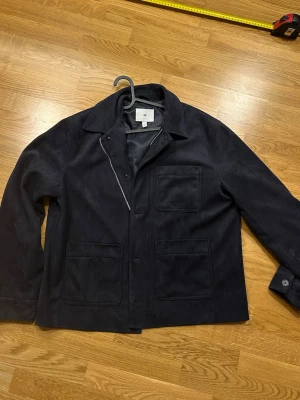 Marinblå overshirt från H&M i mocka  - Stilren mockajacka (mockaimitation) från H&M i mörkblå/svart ton. Mjukt mockaliknande material som ger ett uppklätt men avslappnat uttryck – perfekt till både vardag och kväll. 	•	Storlek L 	•	Normal passform 	•	Mocka/mockaimitation (mjuk yta) 	•	Krage 	•	Dragkedja + knäppning framtill 	•	Bröstficka + två framfickor 	•	Mycket fint skick  Passar perfekt som vår- och höstjacka, och funkar även snyggt över hoodie eller tunn stickad tröja (se outfitbild). 