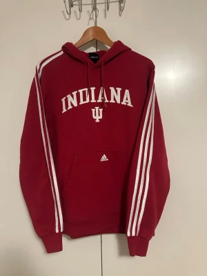 Röd Indiana hoodie från Adidas - Snygg röd hoodie från Adidas med vita ränder längs ärmarna och stor 'INDIANA' text på bröstet. Klassisk huva med dragsko och magficka framtill. Perfekt för dig som gillar college-stil och sportiga detaljer.