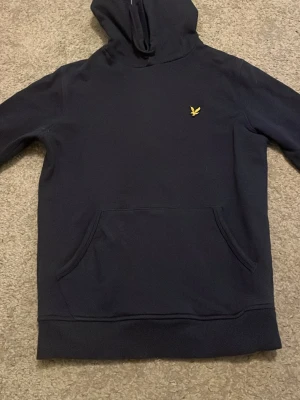 Mörkblå hoodie från Lyle & Scott - Snygg mörkblå hoodie från Lyle & Scott med klassisk gul logga på bröstet. Hoodien har en stor känguruficka framtill och en skön huva. Perfekt för dig som gillar stilrena och bekväma plagg. Materialet känns mjukt och håller värmen bra. Priset går att diskutera!