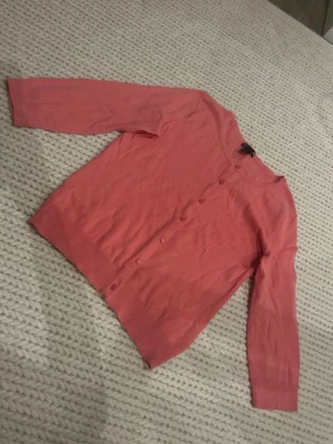 Korallfärgad kofta från H&M - Snygg korallfärgad kofta från H&M med rund halsringning och knappar framtill. Lätt och tunn i materialet, perfekt för lager på lager. Långa trekvartsärmar och ribbade muddar vid ärmslut och nederkant. Enkel och stilren design som passar till mycket.