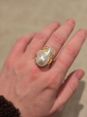 Statementring med stor pärldetalj - Unik ring med en stor, oregelbunden pärlliknande detalj i vitt, infattad i guldfärgad metall. Ringen har en chunky design som verkligen drar blickarna till sig. Perfekt för dig som gillar att sticka ut med accessoarer. Justerbar storlek 