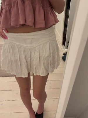 Vit plisserad skort med volanger - Säljer en vit skort med plisserad design och bred linning. Skorten har volangkant och är gjord i ett lätt, luftigt tyg som ger en avslappnad och trendig look. Perfekt för dig som gillar en feminin och bekväm stil.