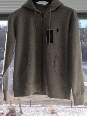 Grå hoodie från Polo Ralph Lauren - Snygg grå hoodie från R Lmed dragkedja framtill, två fickor och klassisk huva med dragsko. Ikoniska logo broderad på bröstet. Tillverkad i mjukt bomullsmaterial som känns skönt mot huden. Perfekt för en avslappnad och stilren look.
