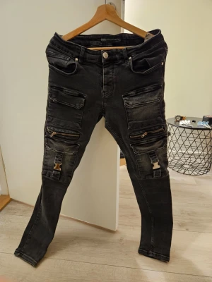 Svarta cargojeans med dragkedjor - Säljer ett par svarta cargojeans med flera stora fickor, dragkedjor och coola spännen på benen. Jeansen har slim fit-modell och är gjorda i stretchigt denimtyg. 