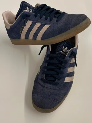 Adidas Gazelle blå/beige sneakers - Snygga Adidas Gazelle sneakers i mörkblå mocka. Skorna har beige häl och gummisula samt mörkblå snörning. Perfekta för dig som gillar retrostil och streetwear. Hör av dig om du har några funderingar eller vill ha fler bilder. Pris kan såklart diskuteras vid en snabb affär.😊