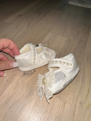 Vita babyskor med pärlor och spetsrosett - Söta vita babyskor i satinliknande material med dekorativa pärlor på remmen och en stor spetsrosett framtill. Skorna har kardborreknäppning och mjuk sula, perfekta för speciella tillfällen som dop eller fest.