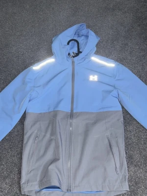 Blå och grå vindjacka från Under Armour - Snygg tvåfärgad vindjacka från Under Armour i ljusblått och grått. Jackan har huva, dragkedja framtill, två sidofickor med dragkedja och reflekterande detaljer på axlar och bröst. Perfekt för blåsiga dagar.