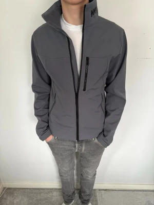 Grå vindjacka från Helly Hansen - Snygg grå vindjacka från Helly Hansen med hög krage och hel dragkedja framtill. Jackan har en bröstficka med dragkedja och två sidofickor. Tillverkad i ett slätt syntetmaterial och har en normal passform. Perfekt för blåsiga dagar.