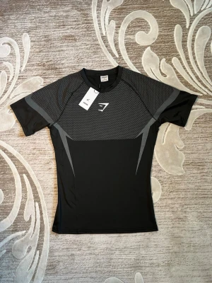 Svart träningströja från Gymshark - Snygg svart träningströja från Gymshark med gråa detaljer och mönster över bröstet. Tillverkad i ett stretchigt funktionsmaterial som andas, perfekt för gymmet eller löpning. Kort ärm och tight passform med logga på bröstet.