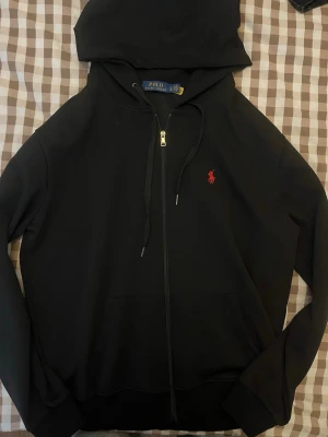 Svart hoodie från Polo Ralph Lauren - Svart hoodie med dragkedja från Polo Ralph Lauren. Klassisk design med huva och dragsnören, samt röd broderad logga på bröstet. Tillverkad i mjukt bomullsmaterial och har känguruficka framtill. Perfekt för en chill och stilren look.