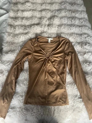 Beige långärmad topp med ringdetalj - Snygg beige långärmad topp med v-ringning och en cool ringdetalj framtill. Figurnära passform och mjukt, stretchigt material som känns skönt mot huden. Perfekt att styla med jeans eller kjol för en trendig look.