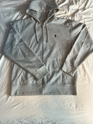 Grå hoodie från Polo Ralph Lauren - Snygg grå hoodie från Polo Ralph Lauren med dragkedja framtill och klassisk huva med dragsko. Tröjan har två fickor på magen och den ikoniska lilla broderade loggan på bröstet. Tillverkad i mjukt bomullsmaterial för skön känsla. Storlek L. Hör av er om ni har frågor!