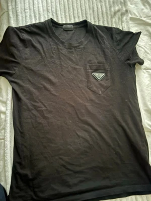 Svart t-shirt från Prada med ficka - Snygg svart t-shirt från Prada med klassisk passform och en bröstficka med den ikoniska Prada-loggan. Perfekt för dig som gillar stilrena och exklusiva plagg. T-shirten har rund hals och korta ärmar.  L storlek snabb äffar 