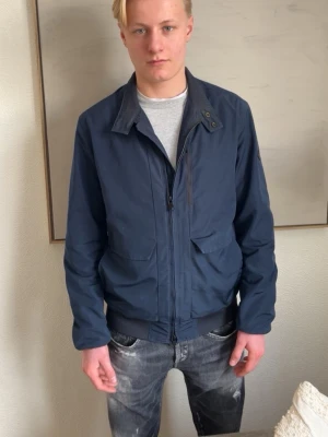  J.Lindeberg Jacka  - Säljer en stilren mörkblå vindjacka från J.Lindeberg. Jackan har dragkedja framtill, stora fickor med lock och en diskret logga på ärmen. Perfekt för blåsiga dagar och enkel att matcha med olika outfits. Klassisk krage och ribbade muddar vid ärmslut.