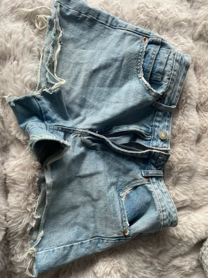 Ljusblå denimshorts med rå kant - Säljer ett par ljusblå denimshorts från H&M med råa, fransiga kanter och klassisk femficksdesign. Shortsen har normal passform och är tillverkade i bomull, perfekta för varma dagar. Stängs med dragkedja och knapp framtill.