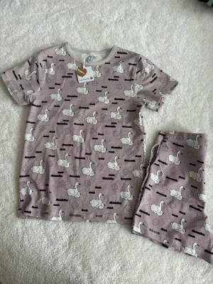 Lila pyjamas med svanmönster - Supersöt tvådelad pyjamas i lila med vita svanar och svarta detaljer. Setet består av en kortärmad tröja och matchande shorts. Tillverkad i mjukt och skönt material, perfekt för mysiga kvällar hemma. Ordinarie pris 499 