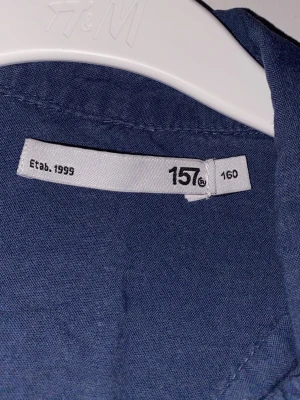 Mörkblå skjorta från H&M - Säljer en stilren mörkblå skjorta från H&M i storlek 160. Skjortan har klassisk krage, knappar framtill och långa ärmar. Perfekt för dig som gillar enkel och tidlös stil.