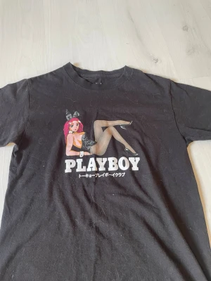 Svart Playboy t-shirt med tryck - Cool svart t-shirt från Playboy med stort tryck av en tecknad figur med rosa hår och texten PLAYBOY på framsidan. T-shirten är i bomull och har klassisk passform med korta ärmar. Perfekt för dig som gillar streetwear och statement-plagg.