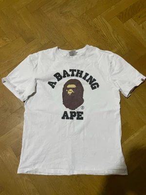 Vit BAPE t-shirt med logga - Säljer en vit t-shirt från A Bathing Ape med den klassiska BAPE-loggan och apans ansikte tryckt på bröstet. T-shirten är kortärmad och har en avslappnad passform, perfekt för en streetwear-look.