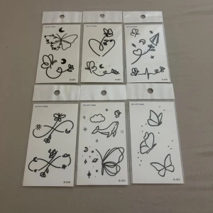 Svarta temporära tatueringar fjärilar - Sju ark med svarta temporära tatueringar i minimalistisk stil. Motiven inkluderar fjärilar, blommor, hjärtan, stjärnor och ett flygplan. Perfekta för maskerad, cosplay eller festival. Lätta att applicera och ta bort.