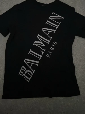 Svart Balmain t-shirt med tryck. PRIS KAN DISKUTERAS!  - Snygg svart t-shirt från Balmain med stort vitt logotyptryck diagonalt över framsidan. Klassisk rund halsringning och korta ärmar. Perfekt för dig som gillar exklusiva streetwear-plagg och vill sticka ut med en ikonisk design.