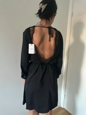 Svart klänning med öppen rygg och spets - Svart kortklänning med lång ärm och markerad midja. Baksidan har en djup öppen rygg med spetsdetaljer och knytband i nacken. Klänningen har en enkel framsida och är tillverkad i ett lätt, mjukt material som ger en snygg passform.