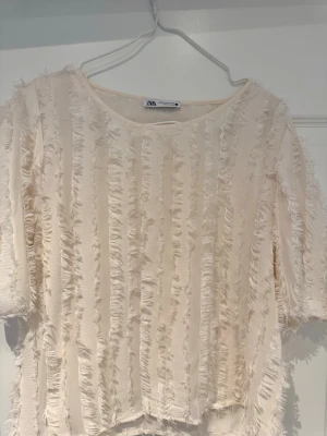 Krämvit fransig topp från Zara - Trendig krämvit topp från Zara med fransiga ränder över hela plagget. Toppen har rund halsringning och trekvartsärmar. Materialet är lätt och ger en cool textur, perfekt för dig som gillar unika detaljer. Litet hål vid armhålan som inte syns.