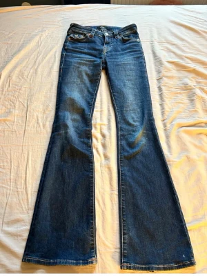 Blå bootcut jeans från True Religion - Säljer ett par blå bootcut jeans från True Religion med klassiska kontrastsömmar och snygga fickdetaljer bak. Jeansen har låg midja och är tillverkade i slitstark denim med lätt tvättad look. Perfekta för dig som gillar retrovibbar och markerade fickor.