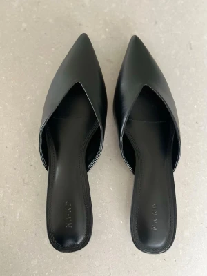 Slip-ins med mini klack  - Säljer ett par svarta ballerinaskor från NA-KD med spetsig tå och öppen häl. Skorna har en stilren design i slätt skinnliknande material och låg klack. Helt oanvända – säljs endast eftersom storleken tyvärr inte passade mig.
