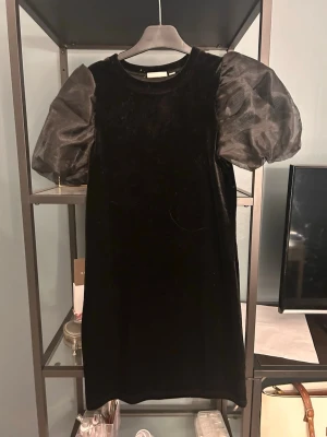 Svart sammetsklänning med puffärm - Stilren svart kortklänning i sammet med stora puffärmar i transparent material. Klänningen har rund halsringning och rak passform, perfekt för dig som vill ha en trendig och elegant look.