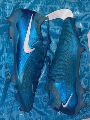 Blå Nike fotbollsskor med vita detaljer - Snygga blå fotbollsskor från Nike med vita swoosh-loggor på sidorna. Skorna har en modern design med struktur på ovandelen och snörning framtill. Tillverkade i syntetmaterial för bra grepp och känsla på planen. Perfekta för dig som vill sticka ut på matchen.