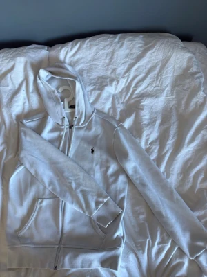 Vit hoodie från Polo Ralph Lauren - Snygg vit hoodie från Polo Ralph Lauren med dragkedja framtill och klassisk huva med dragsko. Ikoniska Polo-loggan broderad på bröstet i svart. Hoodien har två fickor fram och ribbade muddar vid ärmar och nederkant. Perfekt för en clean och stilren look.