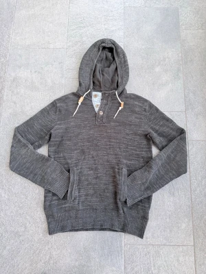 Mörkgrå hoodie med knappar - Mysig mörkgrå hoodie med huva och vita dragsnören med bruna detaljer. Tröjan har knappar vid halsen och en stor magficka framtill. Tillverkad i mjukt stickat material med ribbade muddar vid ärmslut och nederkant. Perfekt för en avslappnad stil.