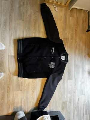 Svart collegejacka med tryck. Vi kan förhandla! - Snygg svart collegejacka från Urban Classics med gråa ärmar och broderade tryck på bröstet. Jackan har ribbade muddar i svart och grått, tryckknappar framtill och klassisk basebollstil. Perfekt för en sportig och avslappnad look.