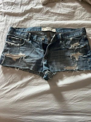  jeansshorts Abercrombie fitch  - Snygga ljusblå jeansshorts från Abercrombie & Fitch med slitna detaljer och råa kanter. Klassisk femficksmodell med låg midja och coola trasiga partier framtill och baktill. Perfekta för en avslappnad och trendig look.