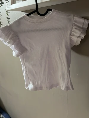 Vit ribbad topp med volangärmar - Supersöt vit topp med ribbad struktur och korta ärmar med volangdetaljer. Perfekt för dig som vill ha en enkel men ändå trendig look. T-shirten har rund halsringning och är tillverkad i mjukt material som känns skönt mot huden.
