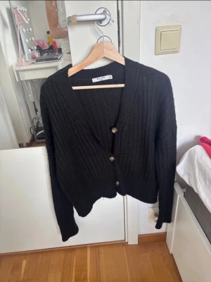 Svart ribbstickad kofta från NA-KD - Svart ribbstickad kofta från NA-KD med v-ringning och tre knappar framtill. Koftan har lång ärm och en lite croppad passform. Perfekt att slänga över en t-shirt eller linne för en chill look.