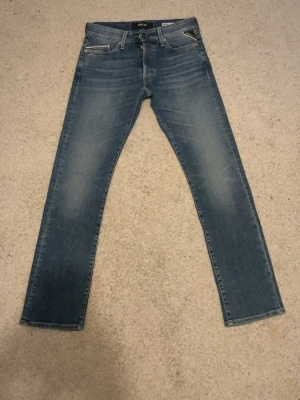 Blå slim fit jeans från Replay - Snygga blå slim fit jeans från Replay med klassisk femficksdesign och diskreta slitningar framtill. Jeansen har normal midja och raka ben. Materialet är denim i bomull med lite stretch för extra komfort. Perfekta för en avslappnad och trendig look. Storlek 29W 32L