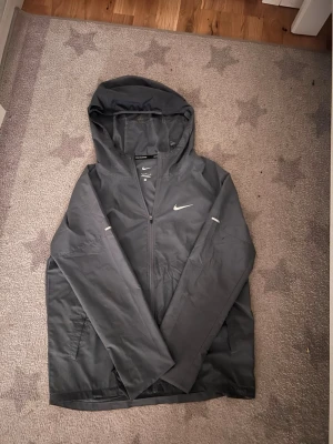 Grå Nike Miller  - Snygg grå vindjacka från Nike med dragkedja och huva. Jackan har diskreta reflexdetaljer på ärmarna och klassisk Nike-logga på bröstet. Perfekt för blåsiga dagar och sportiga outfits. Tillverkad i lätt och smidigt material.